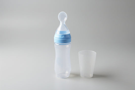 Baby bottle with blue cap and clear cup on a beige surface with green plant in the background